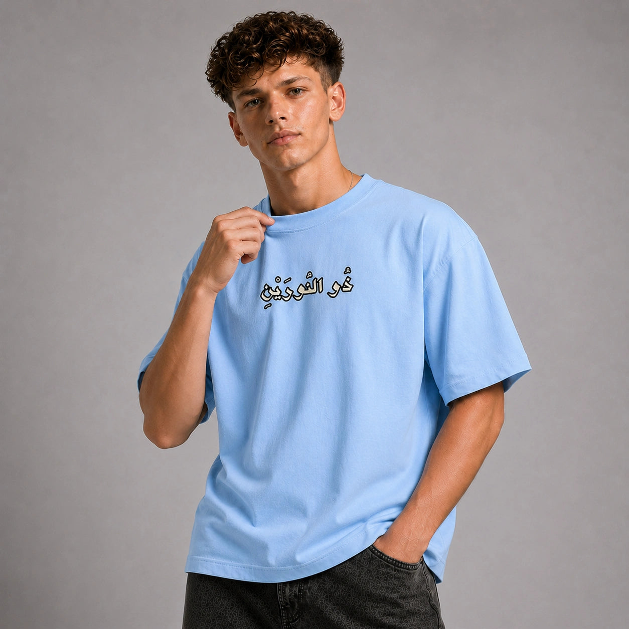 A young man wearing a light blue crewneck t-shirt with black Arabic calligraphy on the chest, posed against a neutral gray studio background.