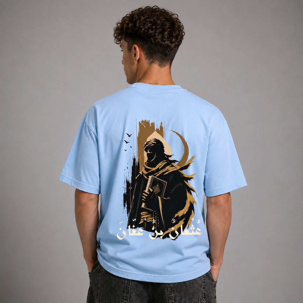 A man viewed from behind wearing a light blue t-shirt with a large graphic print on the back. The graphic features a dark hooded figure holding a sword, a crescent moon, birds, and white Arabic text, set against a plain gray studio background.