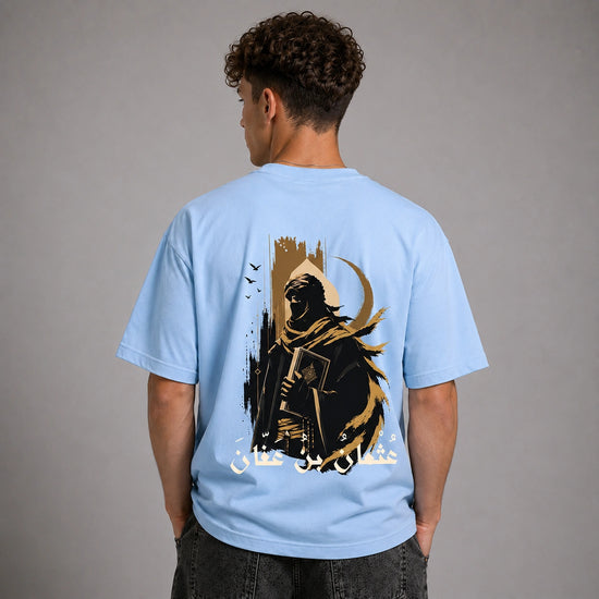 A man viewed from behind wearing a light blue t-shirt with a large graphic print on the back. The graphic features a dark hooded figure holding a sword, a crescent moon, birds, and white Arabic text, set against a plain gray studio background.