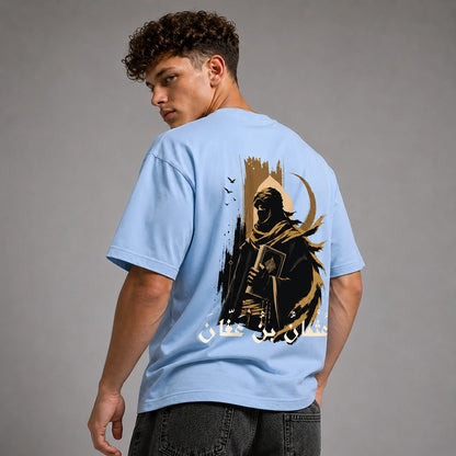 A man wearing a light blue t-shirt with a large black and gold graphic print on the back, photographed against a neutral gray studio background.