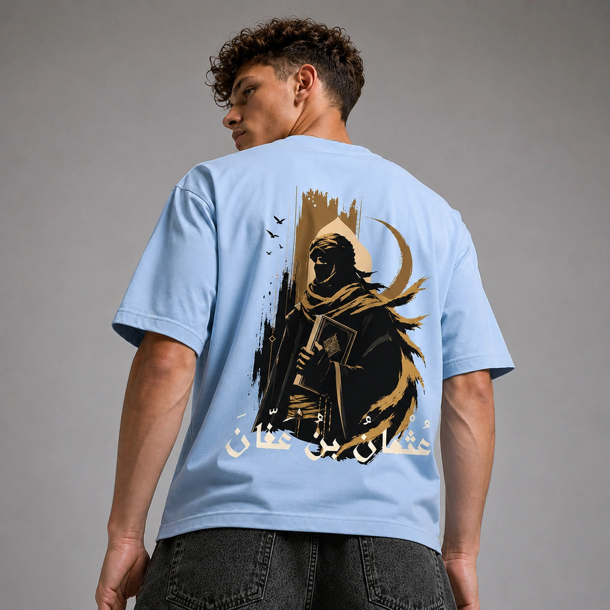 A man wearing a light blue t-shirt, viewed from the back. The shirt features a large graphic print of a hooded figure in black and gold with Arabic calligraphy.