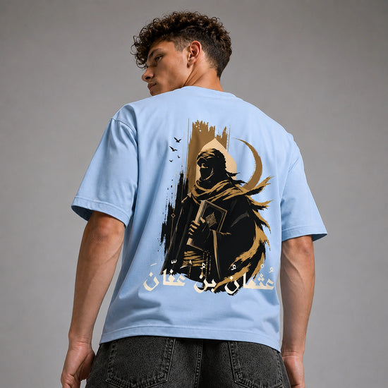 A man wearing a light blue t-shirt, viewed from the back. The shirt features a large graphic print of a hooded figure in black and gold with Arabic calligraphy.