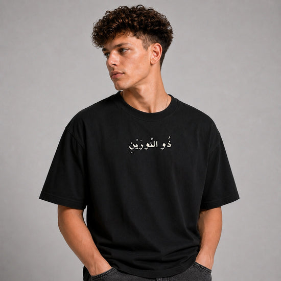 A man wearing a black short-sleeve t-shirt with white Arabic text printed on the chest, posed against a neutral gray studio background.