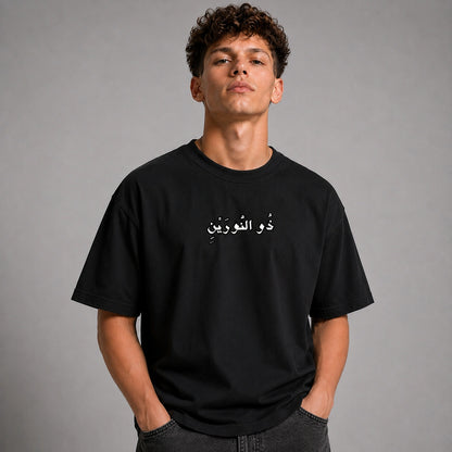 A young man with curly brown hair wearing a black crewneck t-shirt with white Arabic text across the chest, posed against a neutral gray studio background.