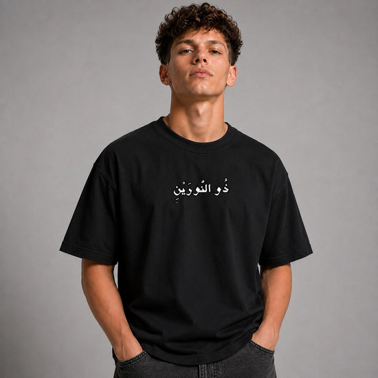 A young man with curly brown hair wearing a black crewneck t-shirt with white Arabic text across the chest, posed against a neutral gray studio background.