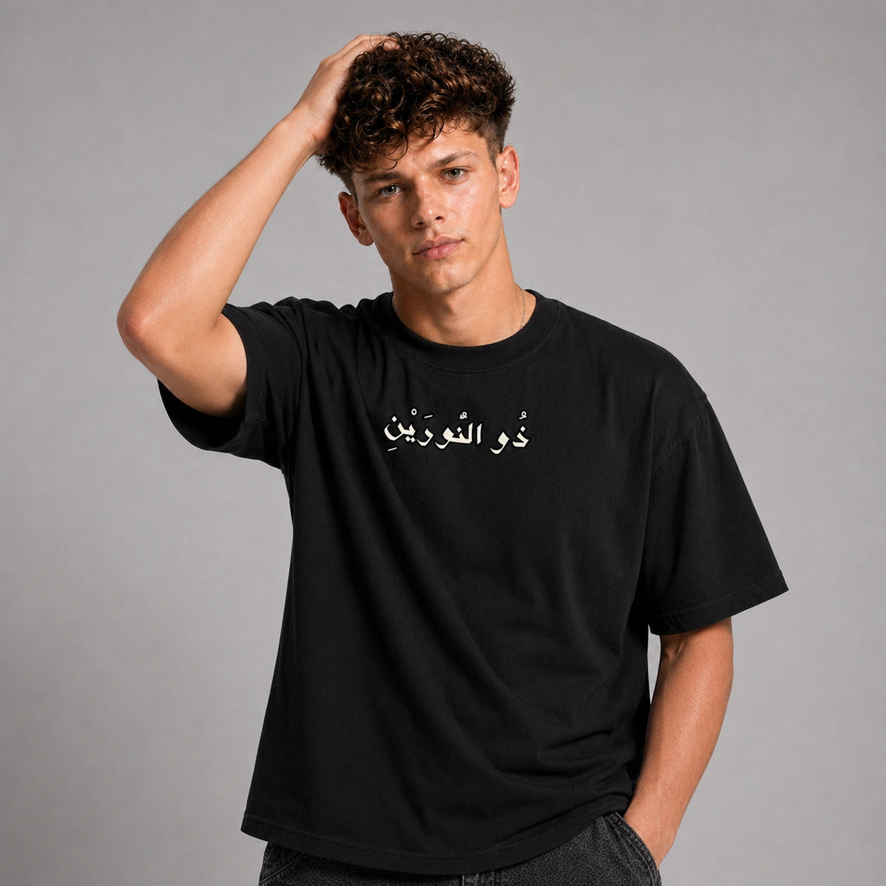 A young man with curly brown hair wearing a black short-sleeve t-shirt with white Arabic calligraphy on the chest, posed against a solid gray studio background.