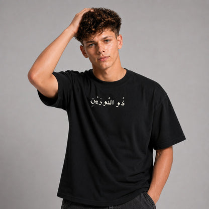 A young man with curly brown hair wearing a black short-sleeve t-shirt with white Arabic calligraphy on the chest, posed against a solid gray studio background.