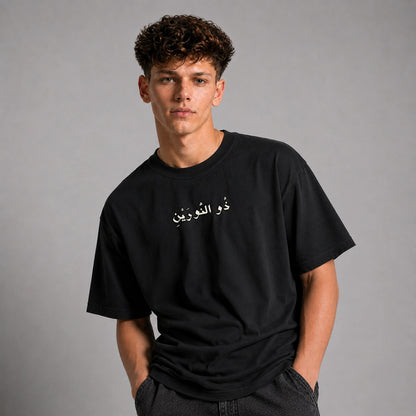 A young man with curly brown hair wearing a black short-sleeve t-shirt with white Arabic calligraphy on the chest, posed against a neutral gray studio background.