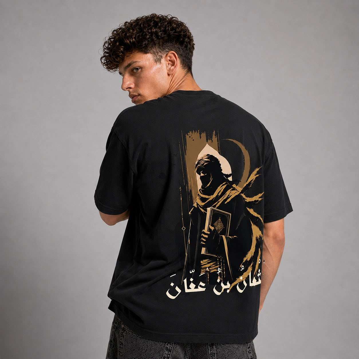 A man wearing a black t-shirt with a large graphic print on the back, photographed from behind against a neutral gray studio background.
