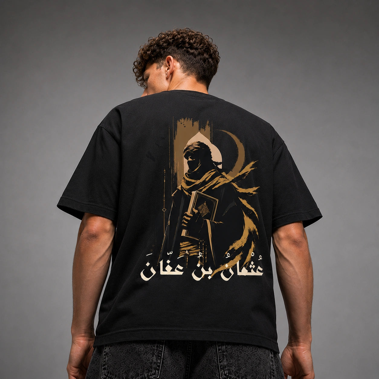 A man viewed from behind wearing a black t-shirt with a large gold graphic print on the back, set against a dark gray studio background.