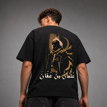 A man viewed from behind wearing a black t-shirt with a large gold graphic print on the back, set against a dark gray studio background.
