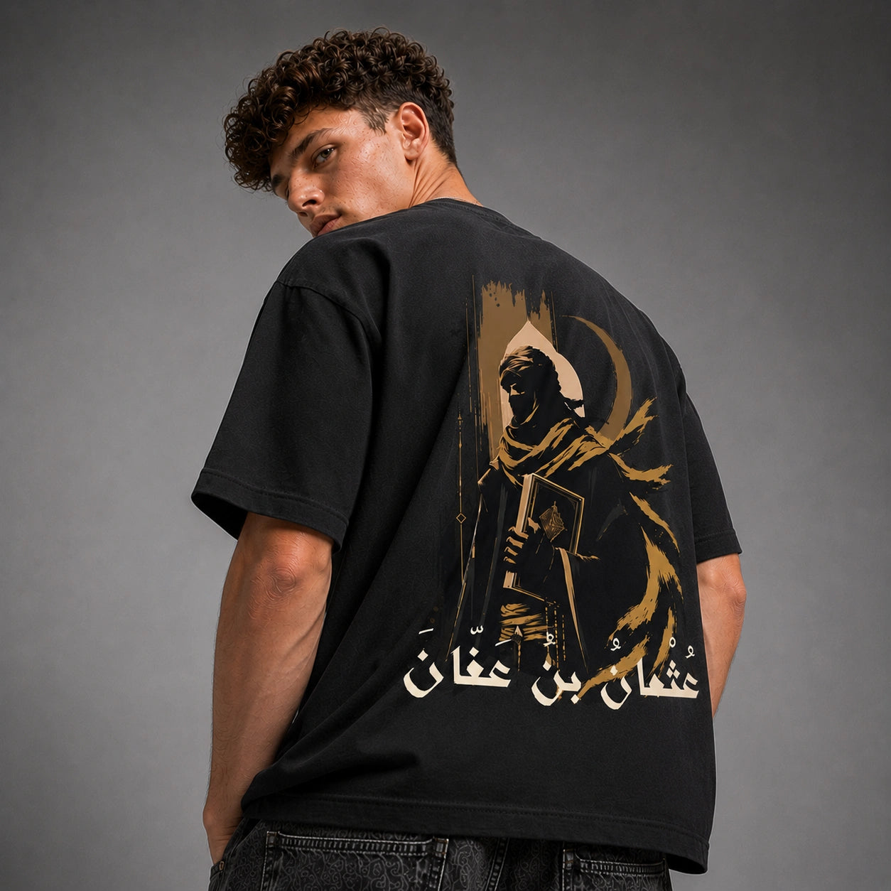A man wearing a black oversized t-shirt with a large graphic print on the back, photographed against a neutral gray studio background.