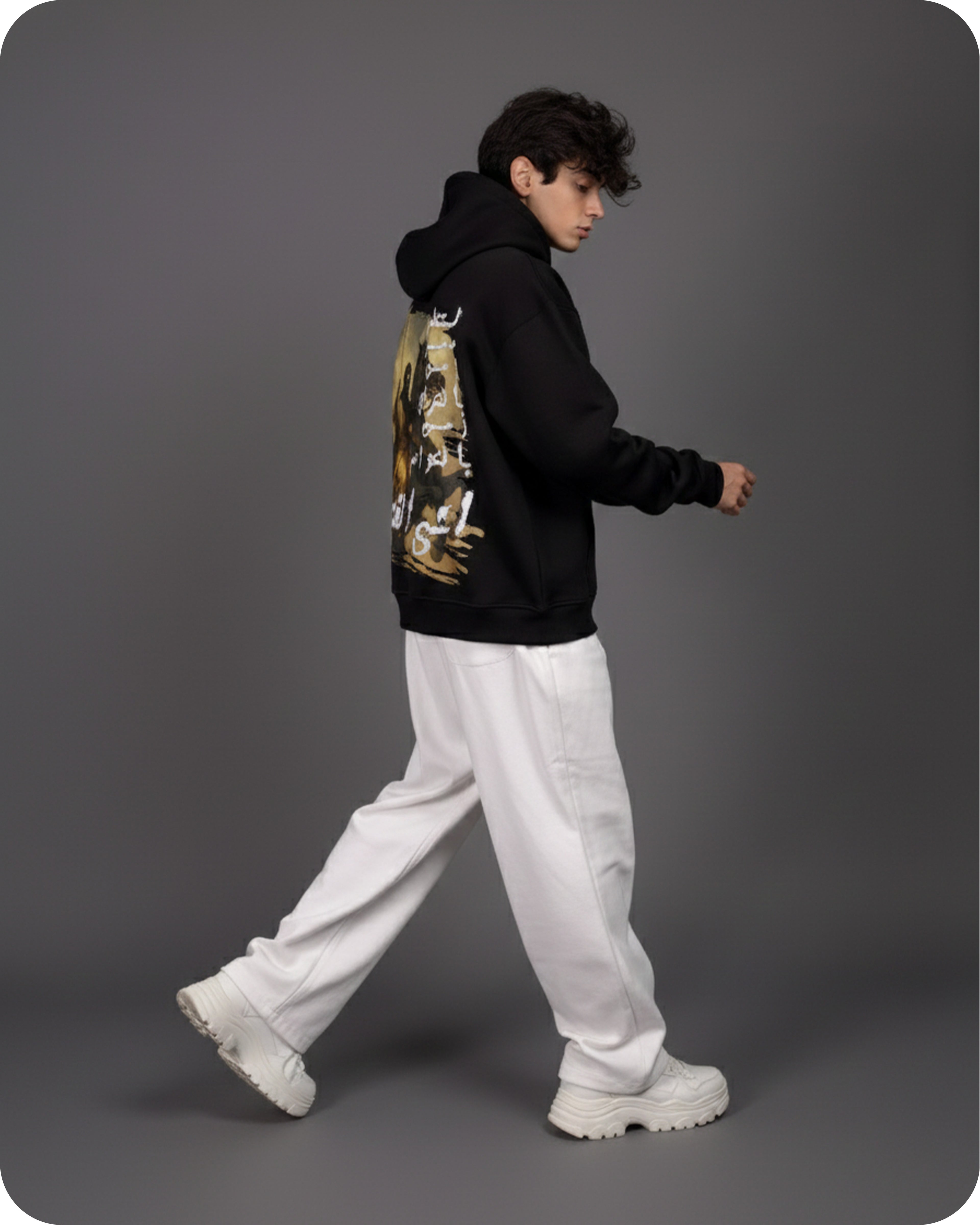 Person wearing a black hoodie with a graphic design, white pants, and white shoes on a gray background