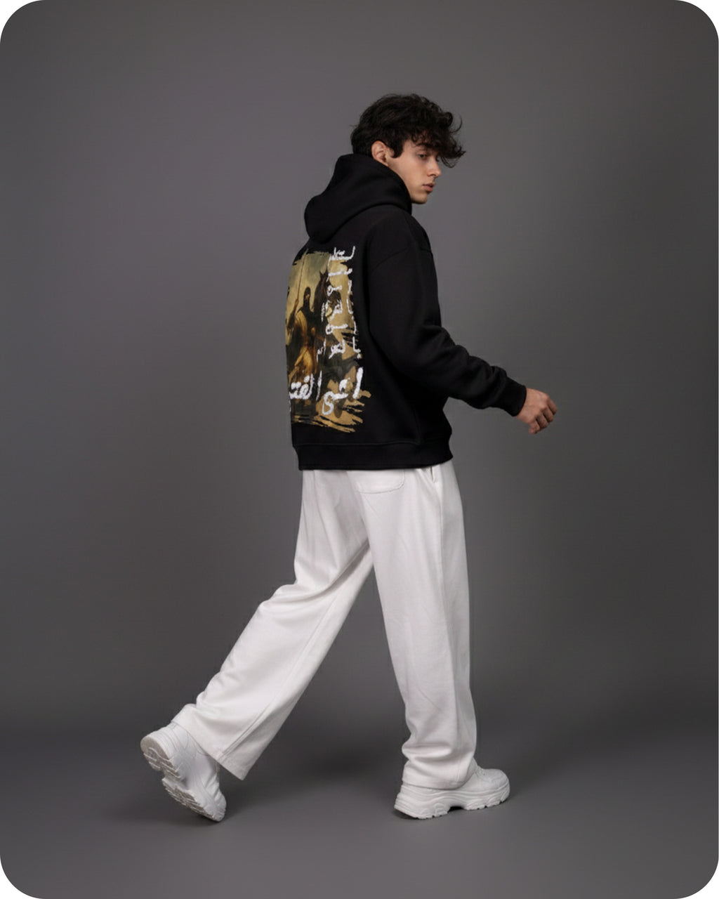 Person wearing a black hoodie with a graphic design and white pants on a gray background