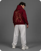 Person wearing a maroon hoodie with a dragon design on the back, standing against a gray background.