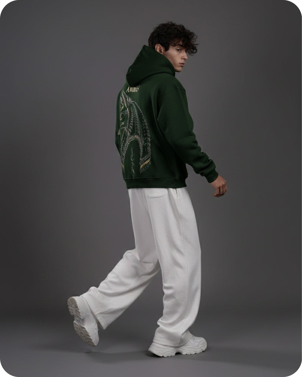 Person wearing a green hoodie with a design on the back, white pants, and white shoes against a gray background
