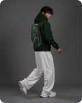 Person wearing a green hoodie with a design on the back, white pants, and white shoes against a gray background