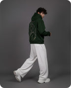 Person wearing a green hoodie with a dragon design on the back, white pants, and white shoes against a gray background.