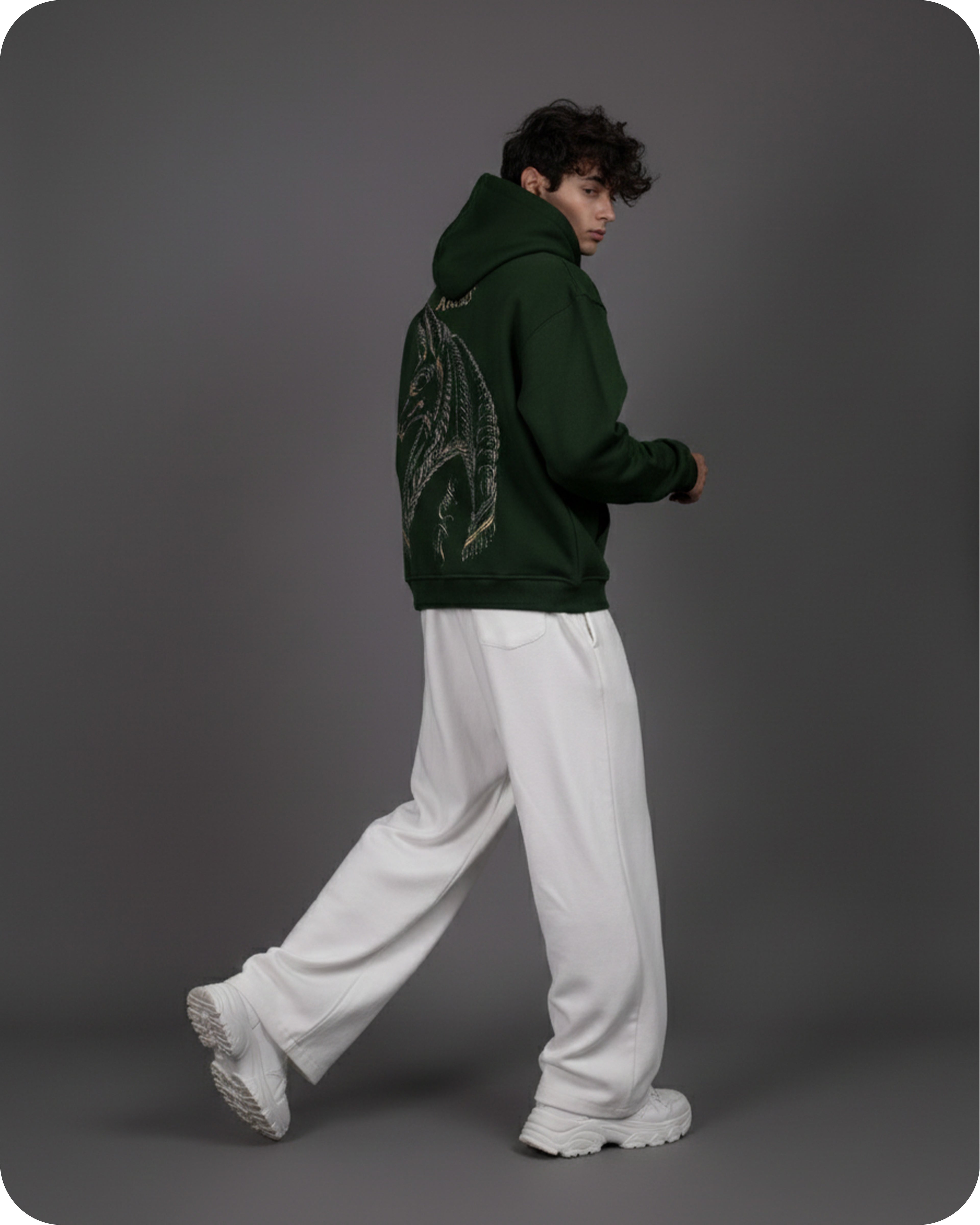 Person wearing a green hoodie with a dragon design on the back, white pants, and white shoes against a gray background.