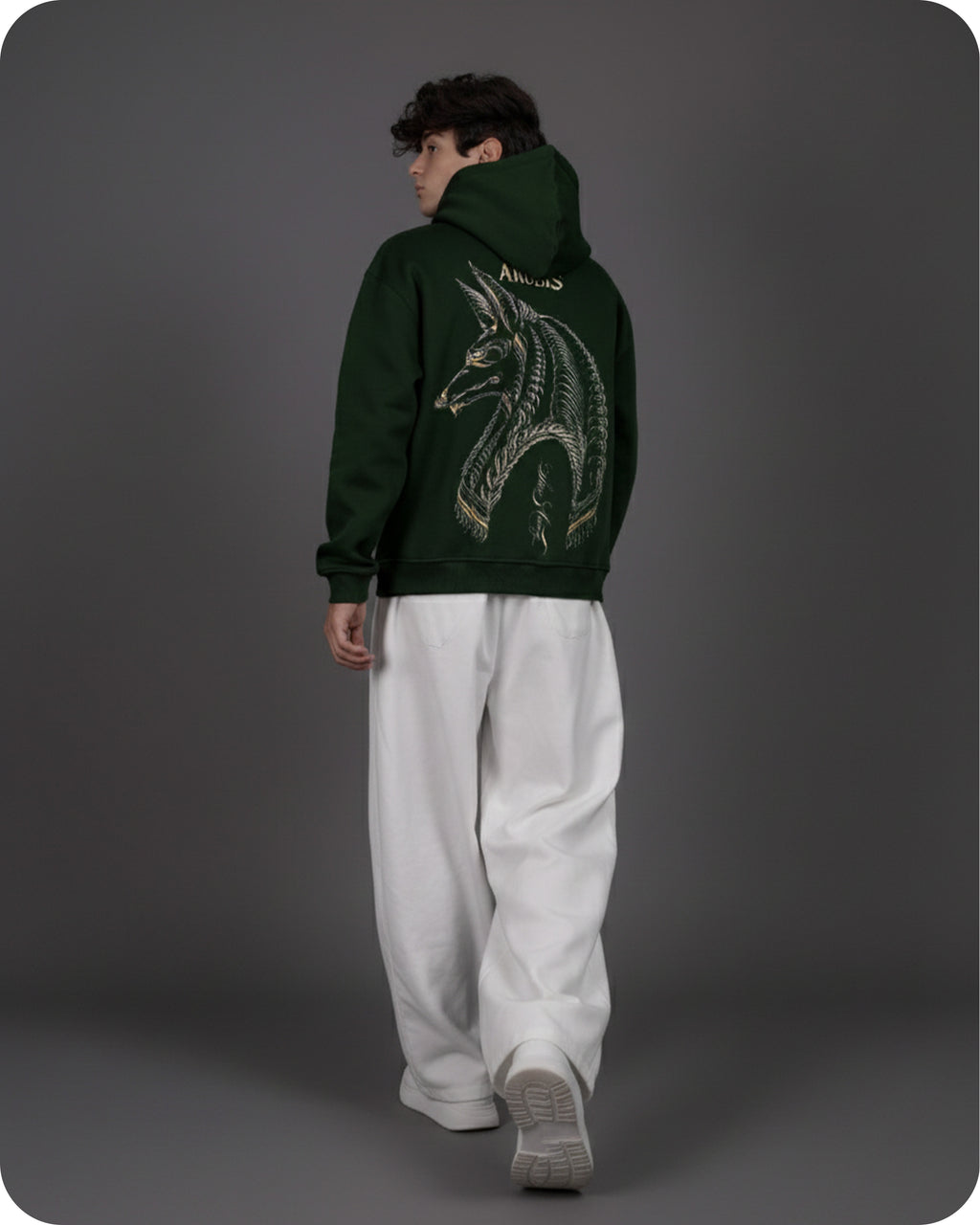 Person wearing a dark green hoodie with a dragon design on the back, standing against a gray background.