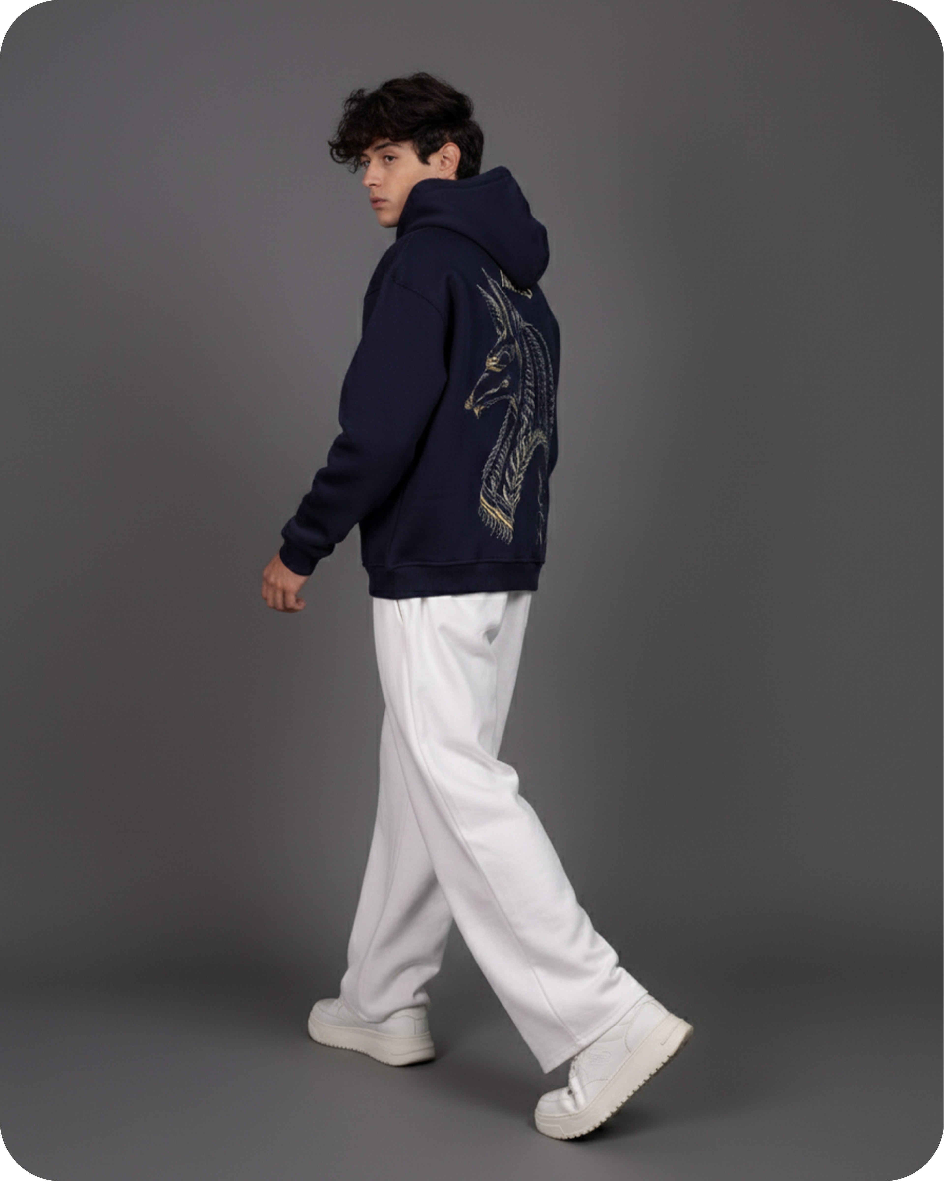 Person wearing a navy hoodie with a dragon design and white pants on a gray background