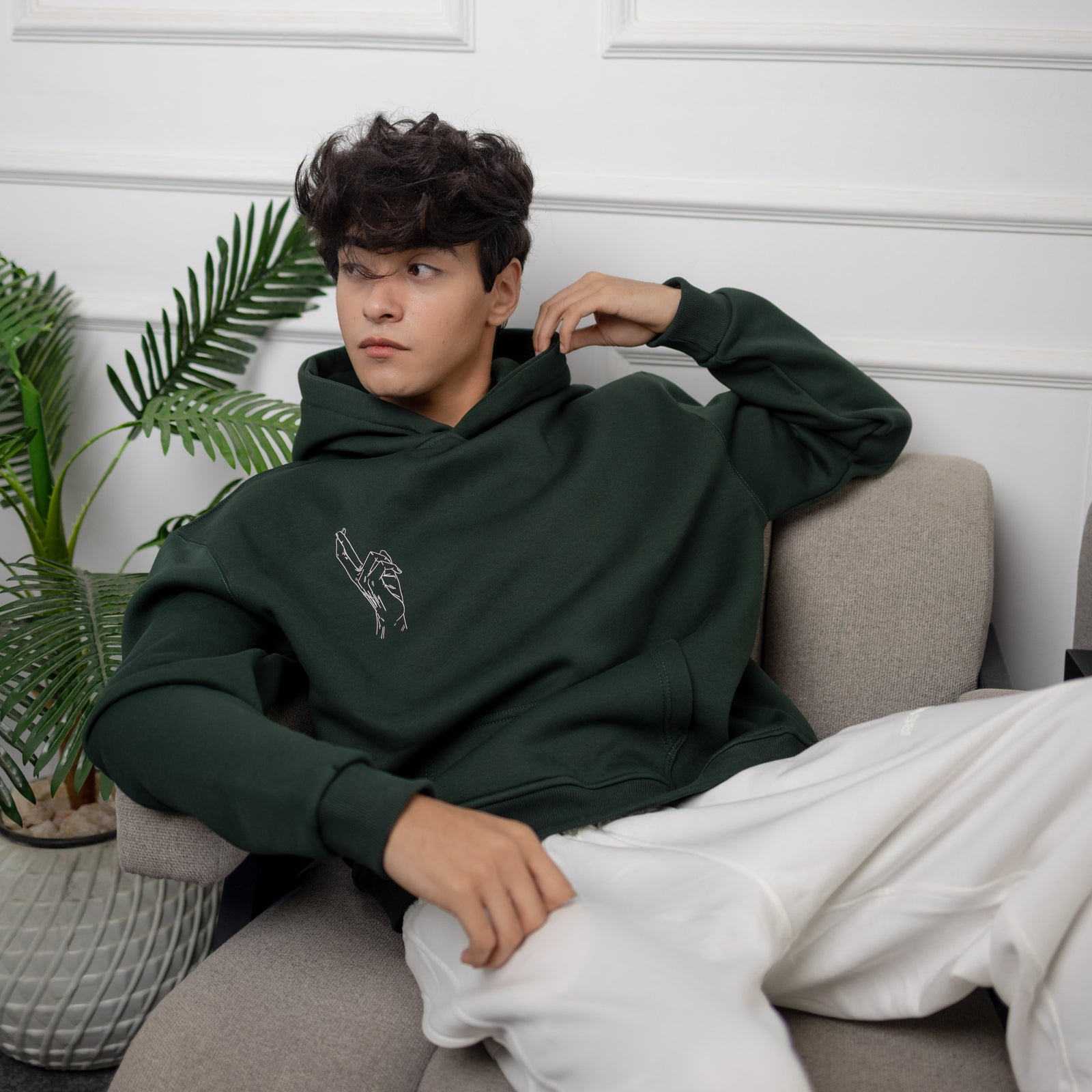 Person wearing a green hoodie sitting on a couch with a white wall and plant in the background