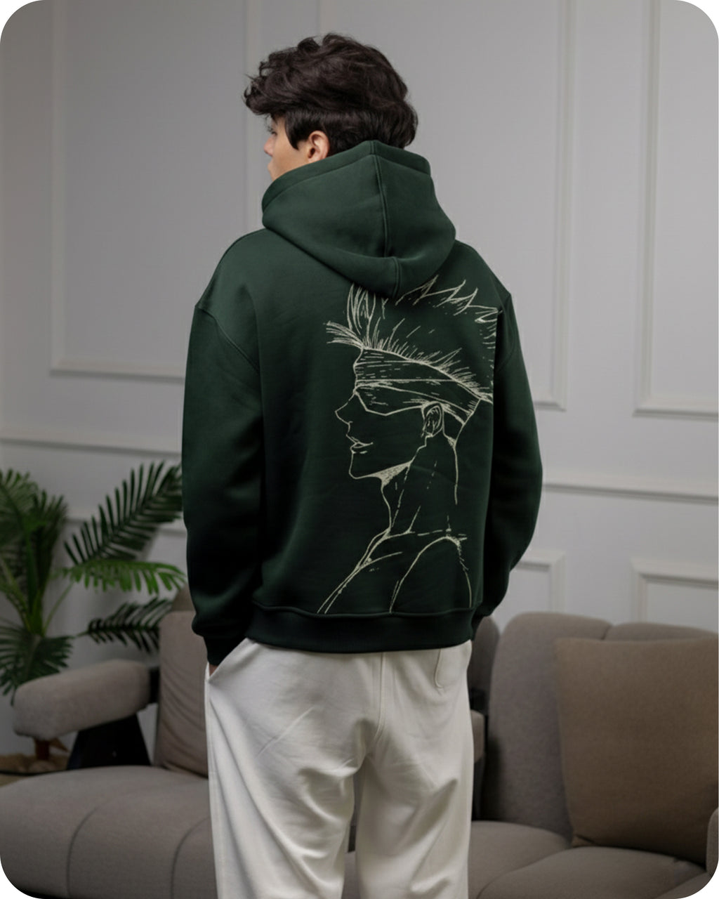 Person wearing a dark green hoodie with a face design on the back, standing in a room with a couch and plant.