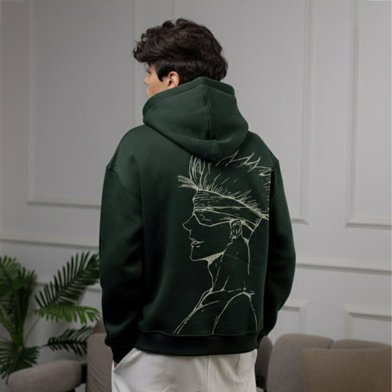 Person wearing a dark green hoodie with a face design on the back, standing in a room with a couch and plant.