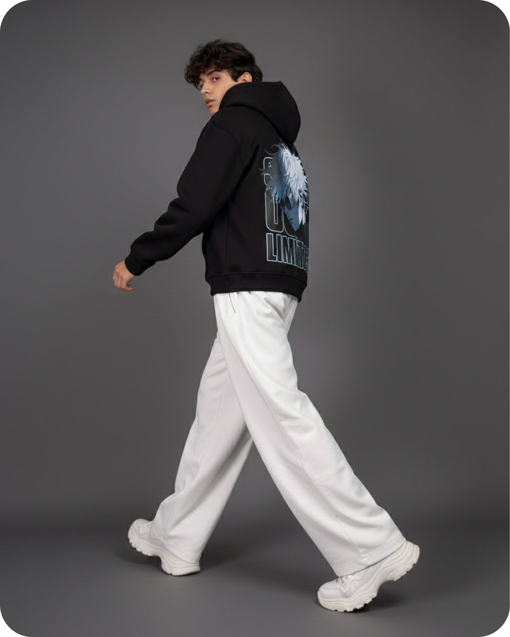 Person wearing a black hoodie with a graphic design on the back, white pants, and white shoes against a gray background