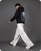 Person wearing a black hoodie with a graphic design on the back, white pants, and white shoes against a gray background