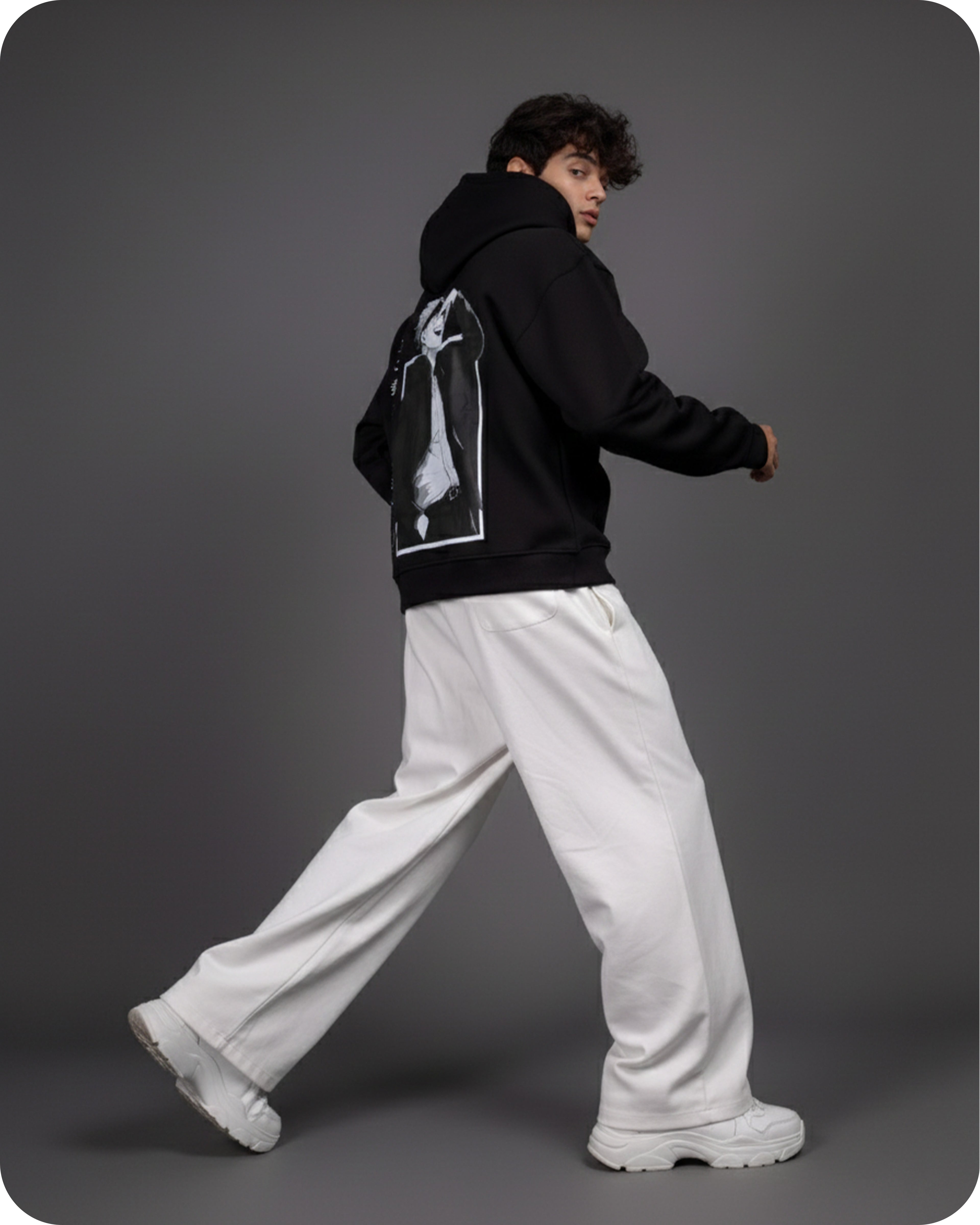 Person wearing a black hoodie with a graphic design, white pants, and white shoes on a gray background