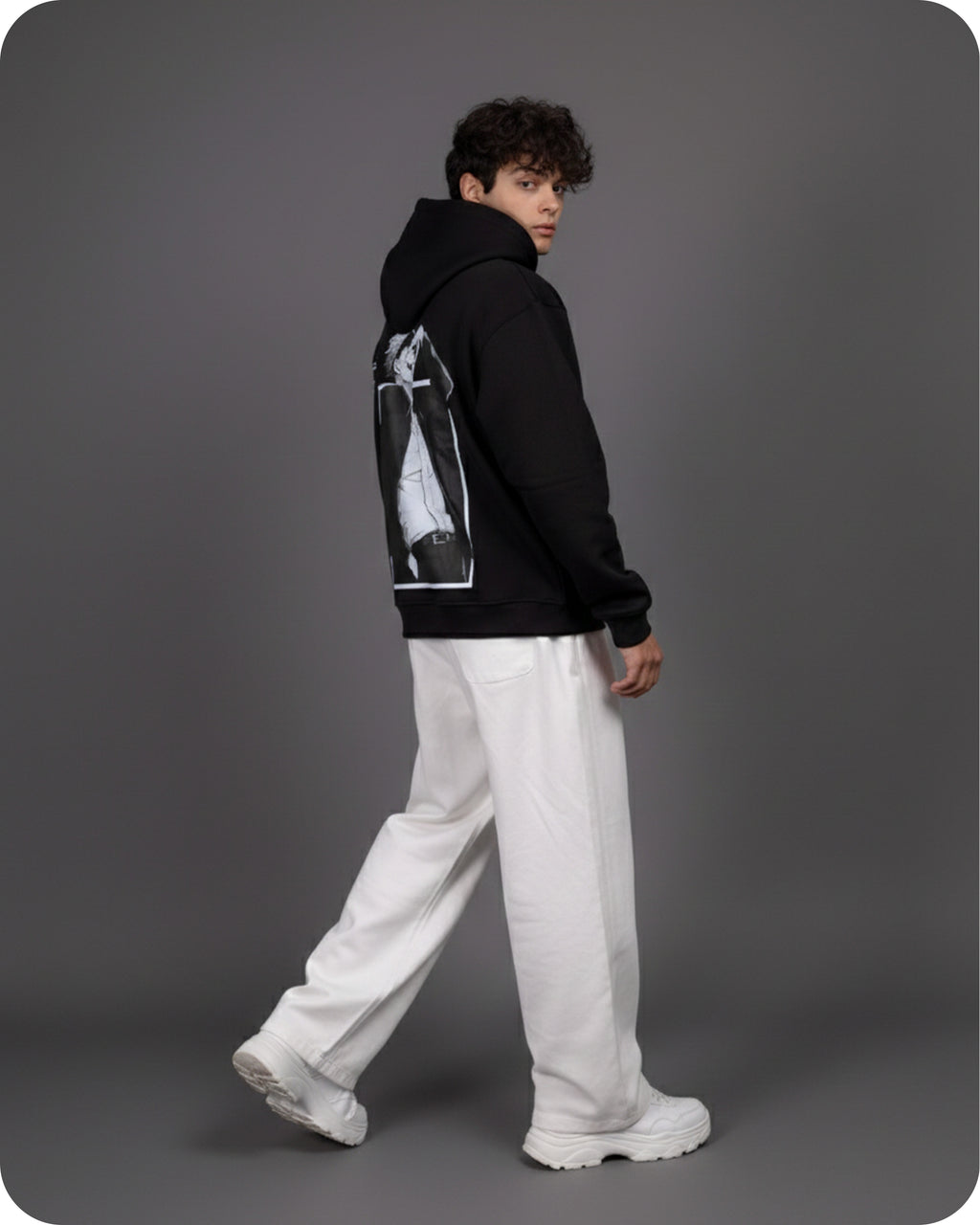 Person wearing a black hoodie with a graphic design on the back, white pants, and white shoes against a gray background