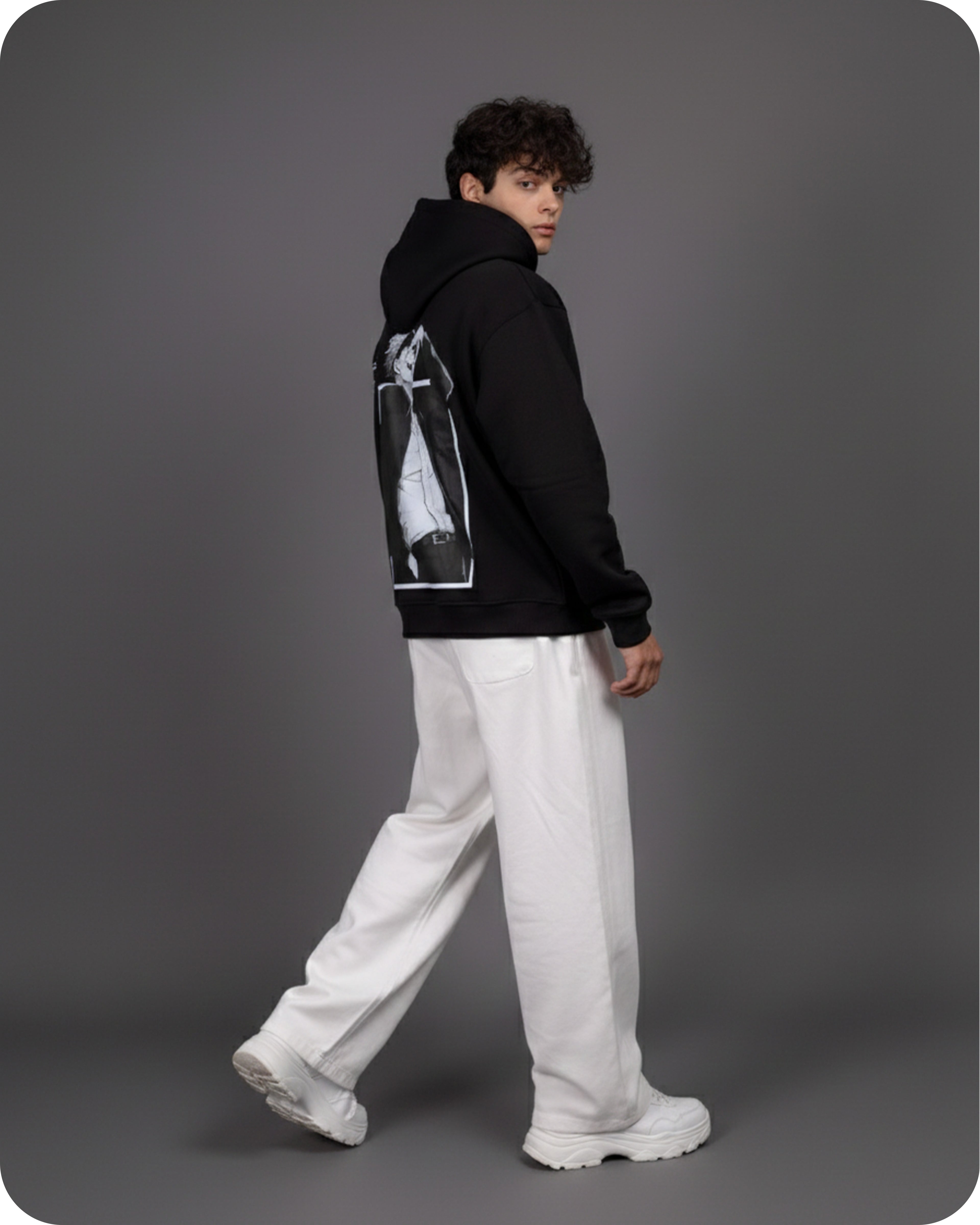 Person wearing a black hoodie with a graphic design on the back, white pants, and white shoes against a gray background