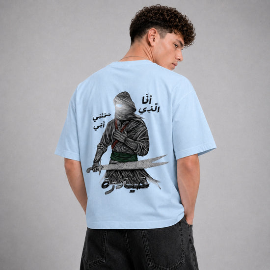 A man wearing a light blue t-shirt, viewed from the back. The shirt features a large graphic print of a hooded figure holding a sword, with Arabic text.