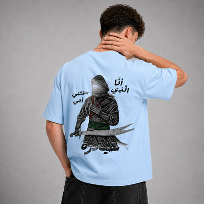 A man shown from the back wearing a light blue t-shirt with a large graphic print of a hooded figure holding a sword and Arabic text. The model is standing against a neutral gray studio background.