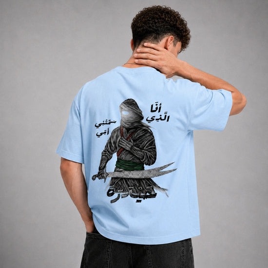 A man shown from the back wearing a light blue t-shirt with a large graphic print of a hooded figure holding a sword and Arabic text. The model is standing against a neutral gray studio background.