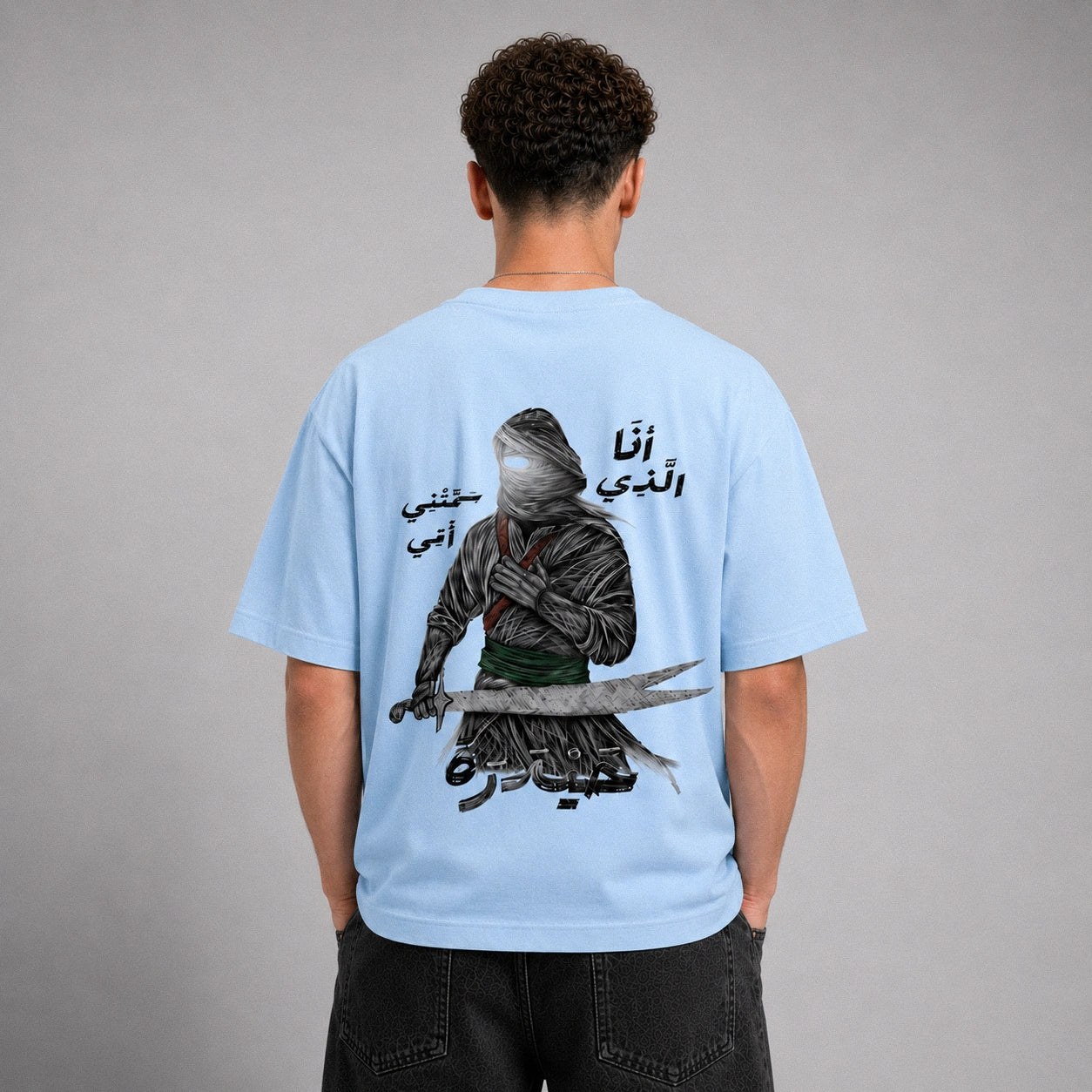 A man viewed from behind wearing a light blue t-shirt with a large graphic print of a hooded figure holding a sword and Arabic text. He is standing against a neutral gray studio background.