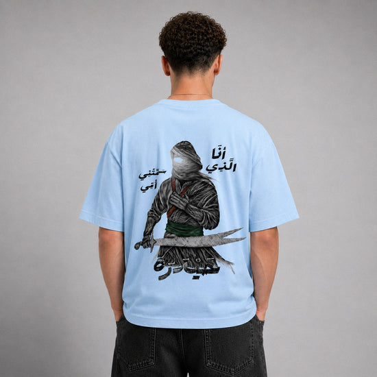 A man viewed from behind wearing a light blue t-shirt with a large graphic print of a hooded figure holding a sword and Arabic text. He is standing against a neutral gray studio background.