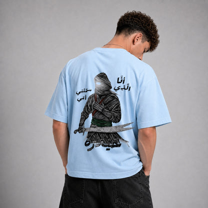 A man wearing a light blue t-shirt, viewed from behind. The back of the shirt features a large graphic print of a hooded figure holding a sword, with black Arabic calligraphy.