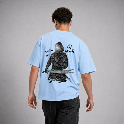 A man viewed from behind wearing a light blue t-shirt with a large graphic print of a hooded warrior holding a sword and Arabic text, paired with dark jeans against a neutral gray background.