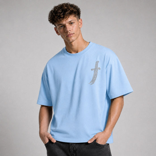 A young man wearing a light blue crewneck t-shirt with a silver glittery sword graphic on the left chest, paired with dark gray jeans, against a neutral gray studio background.