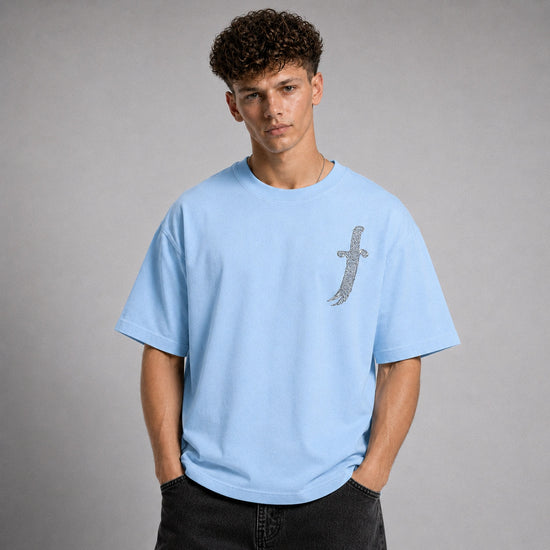 A young man with curly brown hair wearing a light blue crewneck t-shirt with a silver glittery dagger graphic on the left chest, paired with black jeans, standing against a neutral gray studio background.
