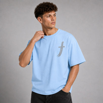 A man wearing a light blue crewneck t-shirt with a silver glittery dagger graphic on the left chest, posed against a solid gray studio background.