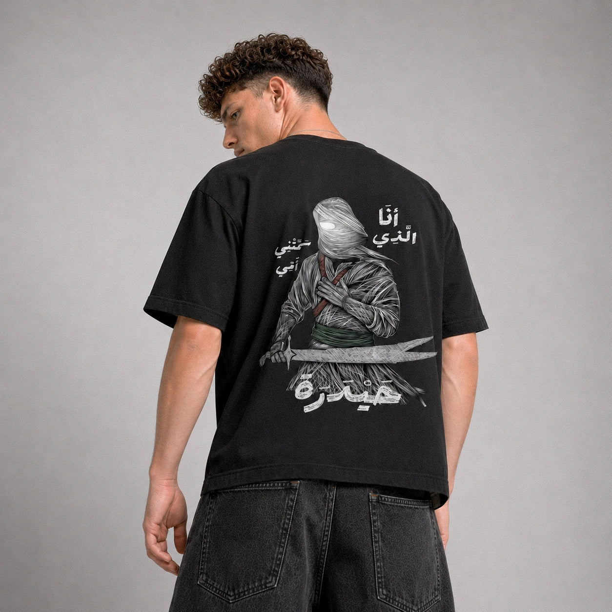 Person wearing a black t-shirt with a graphic design and text on the back, standing against a gray background.