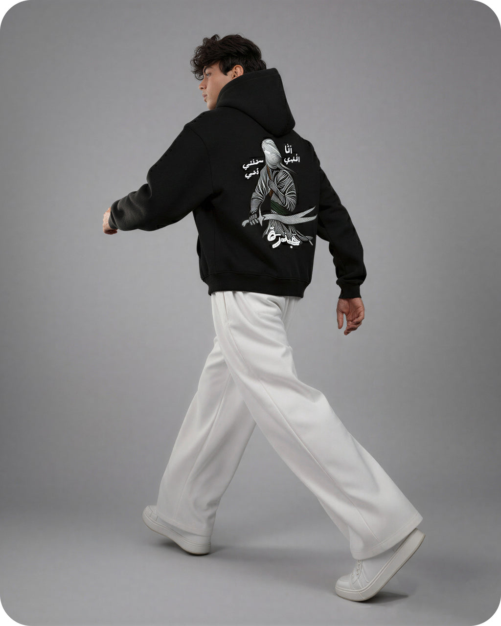 Person wearing a black hoodie with white text and graphics on the back, walking against a gray background
