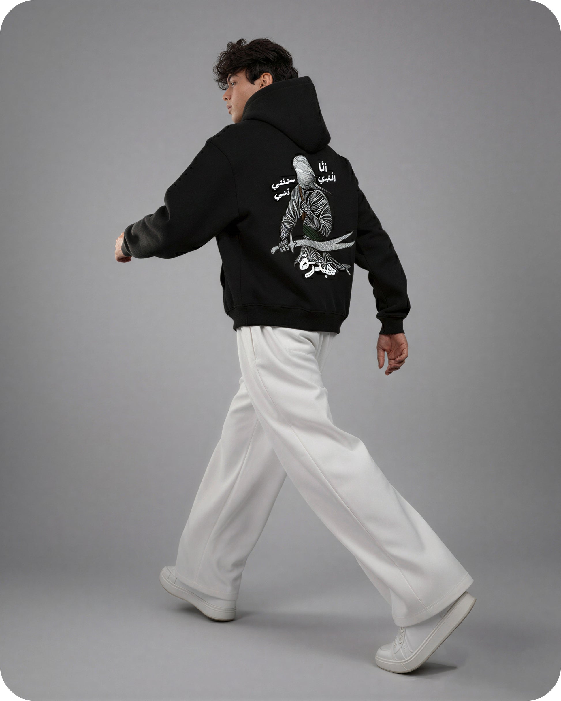 Person wearing a black hoodie with white text and graphics on the back, walking against a gray background