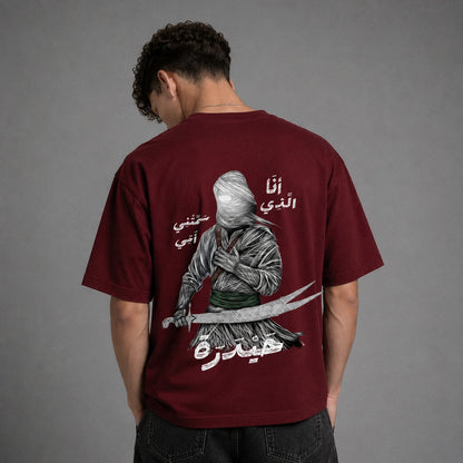 A man wearing a maroon t-shirt from the back, featuring a large graphic print of a bandit with Arabic text. The background is a solid gray studio backdrop.