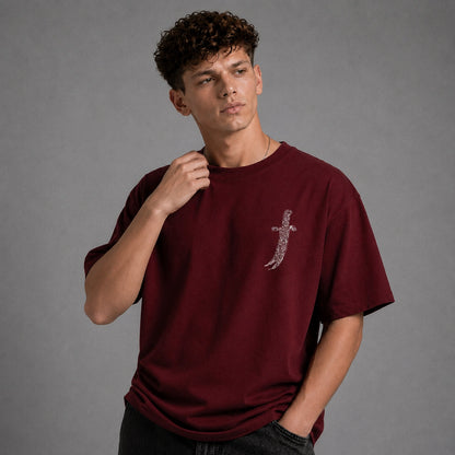 A young man wearing a maroon short-sleeve t-shirt with a silver sword graphic on the left chest, posed against a solid gray studio background.