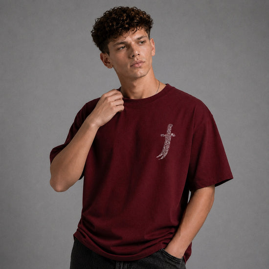 A young man wearing a maroon short-sleeve t-shirt with a silver sword graphic on the left chest, posed against a solid gray studio background.