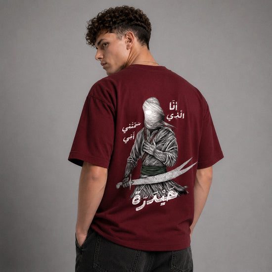 A young man wearing a maroon short-sleeve t-shirt with a distressed silver sword graphic on the left chest, posed against a solid gray studio background.